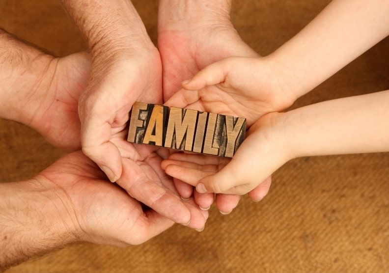Multiple generations of hands holding a wooden block that says family, symbolizing estate planning and family legacy meetings.