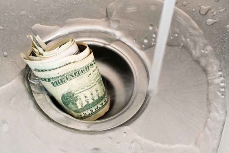 A roll of US dollar bills sitting inside a stainless steel sink drain as water runs around it, illustrating the tax and planning leaks that can erode investment portfolios.